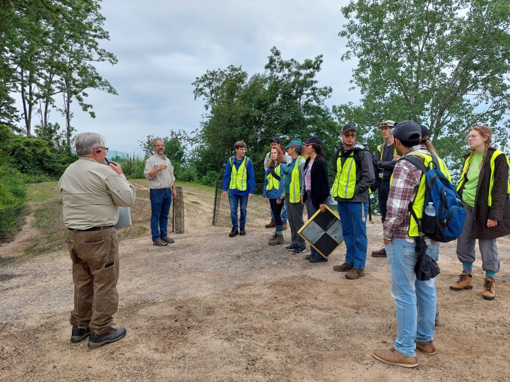 Group of about a dozen people in safety vests standing at a Lake Michigan bluff, listening to the director give instruction