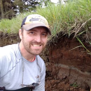 Trent Adams wearing a hat kneeling next to a soil outcrop with tsunami deposit.