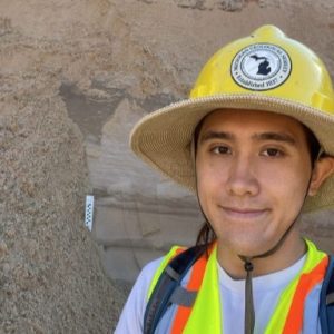 Augustine Lodise wearing a hard hat and safety vest in front of an outcrop of sand.