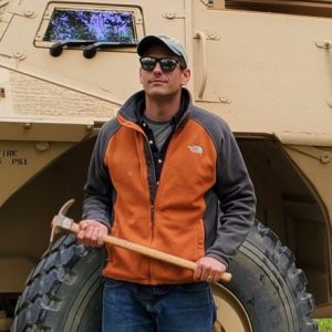 Nate Erber holding a rock hammer in front of a big-wheeled vehicle.