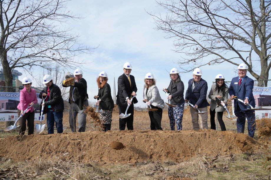 A group of people wearing hard hats stand in a line outdoors, each holding a shovel as soil is being turned during a ceremonial groundbreaking event.