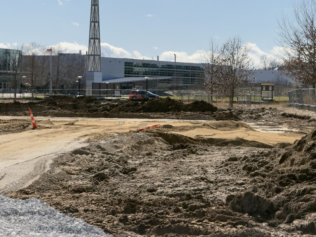 A cleared construction area with dirt and tire tracks in front of a multi‑story brick and glass building.