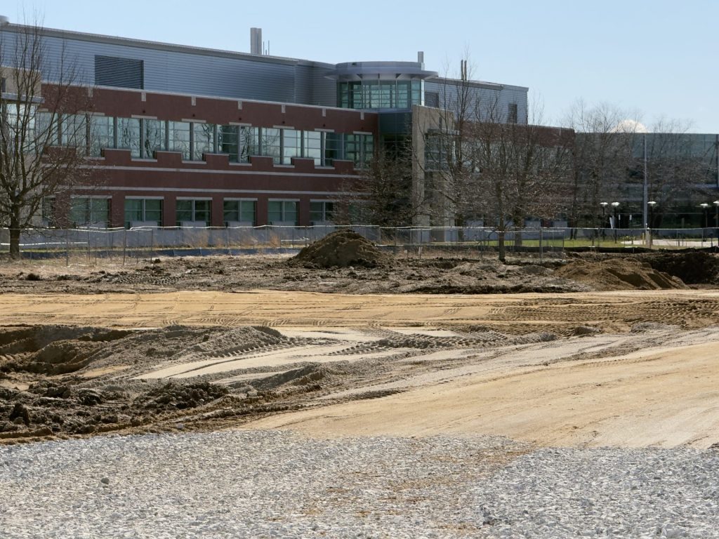 Construction site with dirt ground and tire tracks in front of a large brick building.