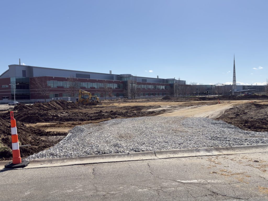 A cleared construction area with dirt and tire tracks in front of a multi‑story brick and glass building.