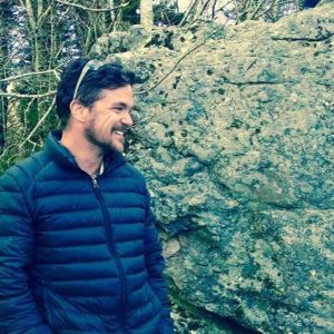 Eric Schuemann wearing sunglasses and a dark colored jacket while standing in front of a rock outcrop.