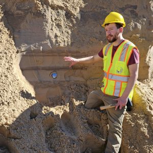 Tom Valachovics wearing a hard hat and safety vest pointing his hand towards an outcrop of sand.