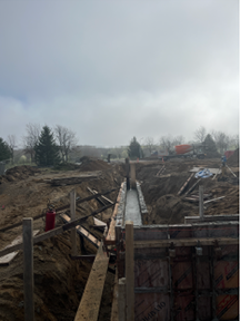 Long, narrow construction trench with wooden formwork running down the center, surrounded by excavated soil and temporary bracing, with construction materials scattered nearby and trees and an overcast sky in the background.
