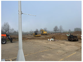 Wide view of a construction site with excavators working on a graded dirt area next to an asphalt parking lot, with construction materials and equipment in the foreground and trees under a clear sky in the background.