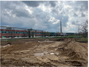 Graded, muddy construction site with tire tracks and shallow excavations in the foreground, with a school building, fencing, and a tall spire visible in the background under cloudy skies.