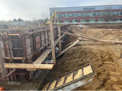 Excavated construction site with a deep trench supported by wooden formwork and bracing, surrounded by sloped sandy soil, with an excavator and a brick building in the background.