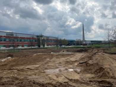 Graded, muddy construction site with tire tracks and shallow excavations in the foreground, with a school building, fencing, and a tall spire visible in the background under cloudy skies.
