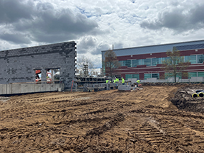 Construction site with a partially built concrete block wall, workers and materials nearby, graded dirt with tire tracks, and a red‑and‑gray building in the background under a cloudy sky.