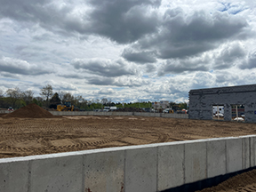 Concrete retaining wall in the foreground with a large dirt construction area, a concrete building structure to the right, and heavy equipment in the distance under a cloudy sky.