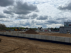 Wide view of an active construction site with tracked dirt, a concrete retaining wall in the foreground, dirt piles and heavy equipment in the midground, and buildings under a cloudy sky.