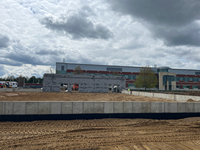 Partially constructed concrete building with a long concrete wall in the foreground, graded dirt surface, and an existing red‑and‑gray building in the background beneath a cloudy sky.