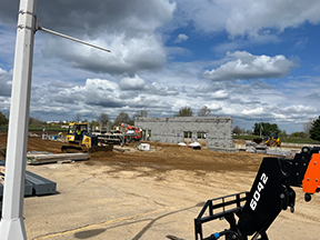 Construction site with a concrete block building under construction, dirt grading in progress, heavy machinery, and construction equipment visible beneath a partly cloudy sky.