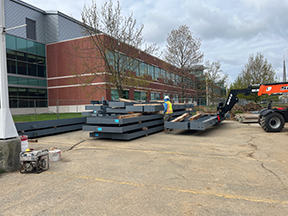 Stacked structural steel components on pavement with construction equipment and a worker nearby, set in front of an existing red‑brick and glass building on an overcast day.