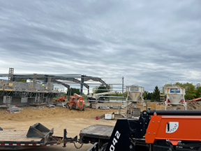 Wide construction site with structural steel framing, a large canopy in progress, and construction equipment on a dirt lot under cloudy skies.