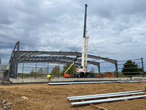 Steel building frame under construction with a crane lifting beams, surrounded by a dirt worksite and stacked metal materials under cloudy skies.