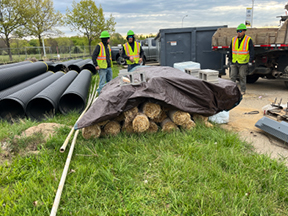 Construction crew standing near stacks of large black pipes and covered materials on grass, with equipment and a truck parked at the site.