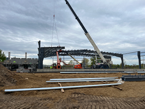 Crane lifting steel beams into place on a large building frame, with workers, stacked metal pieces, and a muddy construction site under overcast skies.