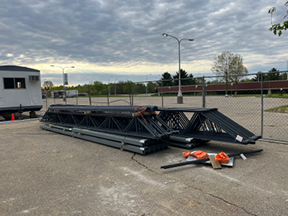 Stacked steel roof trusses and beams stored on a paved surface near a construction site, with a temporary building and fencing in the background.