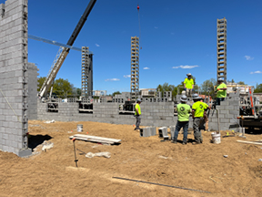 Construction workers installing concrete block walls with vertical column forms and a crane lifting materials, on a dirt construction site under a clear sky.