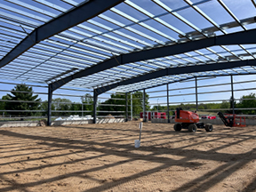 Steel building frame with roof decking installed over a flattened dirt floor, featuring a red boom lift positioned inside the open structure.