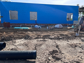 Blue sheathed exterior wall with window openings under construction, surrounded by a dirt worksite with trenches, piping, and scattered building materials.