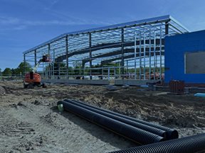 Steel-framed building under construction adjacent to a blue sheathed wall, with an excavator, large black drainage pipes, and a graded dirt site in the foreground.