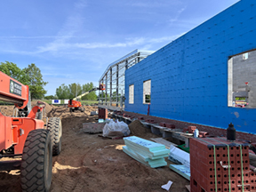 Blue sheathed building wall with multiple window openings under construction, with stacked insulation boards, bricks, and a boom lift along a dirt worksite beside a steel-framed structure.