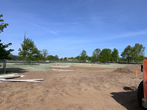 Wide view of a cleared dirt construction site with graded ground, scattered materials, and trees lining the background under a clear blue sky.