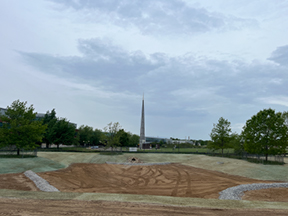 Graded excavation area with curved gravel swales and tire tracks, surrounded by grass and trees, with a tall spire visible in the distance under an overcast sky.