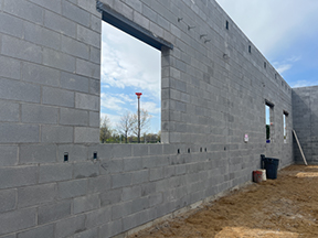 Concrete block wall under construction with large window openings, sandy ground, and tools placed along the base.