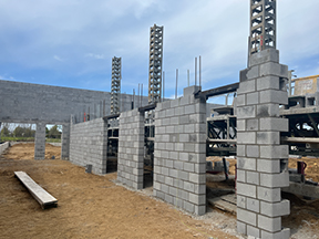 Partially built concrete block structure with multiple wall openings, vertical rebar columns, and interior scaffolding on a dirt construction site.