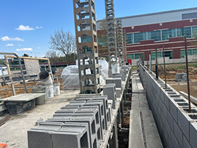 Rows of stacked concrete masonry units and vertical column forms along a partially built wall at a construction site, with rebar and equipment visible.
