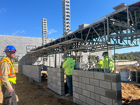 Construction workers laying concrete blocks under elevated scaffolding, with vertical rebar columns and a partially built wall on a dirt site.