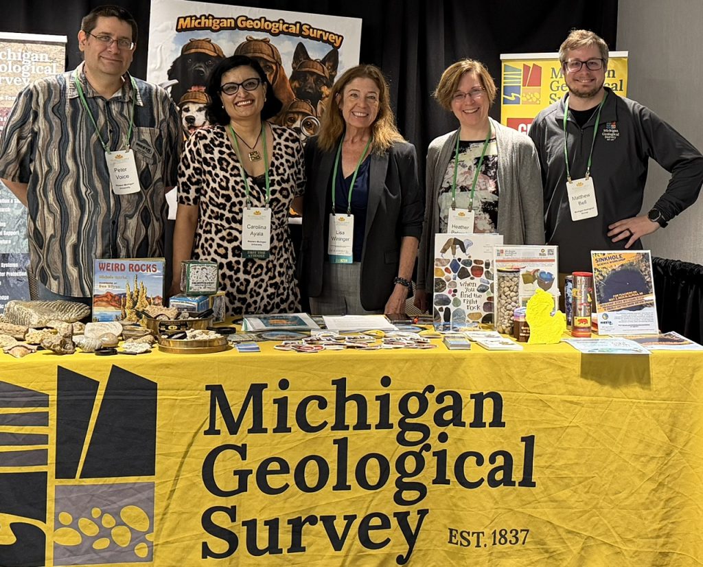 Five people standing behind a Michigan Geological Survey table display with rock samples, books, and educational materials at an indoor outreach event.