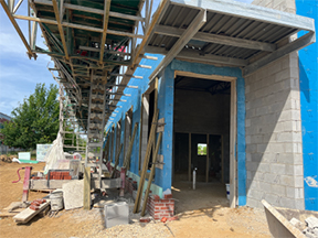 Construction site showing a concrete block building exterior with blue sheathing, scaffolding overhead, and materials and tools scattered along a dirt work area beside an open entry doorway.