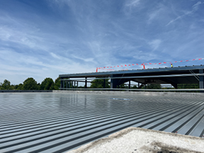 Wide view of a steel-framed structure under construction with roof decking installed, seen from a corrugated metal surface under a partly cloudy sky.