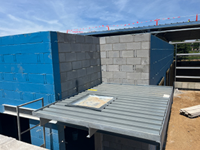 Concrete block and blue sheathed exterior walls forming a corner of a building under construction, with an open roof area, metal canopy below, and construction materials on a dirt ground.