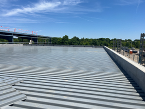 Wide view of corrugated metal roof decking installed on a building under construction, bordered by low concrete walls, with a steel-framed structure and trees in the distance under a blue sky.