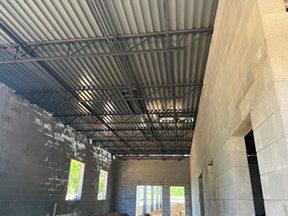 Interior of a concrete block building under construction with corrugated metal roof decking and exposed steel trusses overhead, window openings along one wall, and piles of dirt on the unfinished floor. Provide your feedback on BizChat