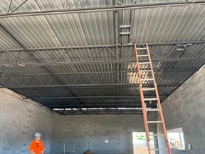 Interior of a concrete block room with corrugated metal roof decking and exposed steel trusses, featuring a tall ladder leaning up to the ceiling and a construction worker standing on the dirt floor below.