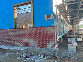 Partially constructed building exterior with a red brick veneer base beneath blue sheathing, an open doorway and window, scaffolding overhead, and construction debris scattered along the dirt ground.
