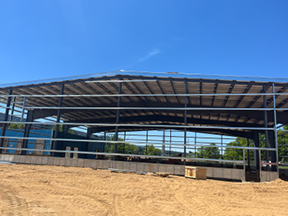 Wide view of a steel-framed building with roof decking installed, open sides, and a sand-covered construction area in the foreground under a clear blue sky.