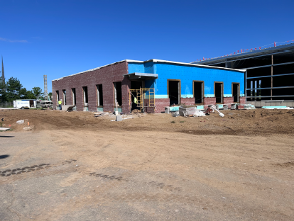 Partially completed building with red brick veneer and blue sheathing, multiple window openings, scaffolding at the entrance, and a wide dirt construction area under a clear blue sky.
