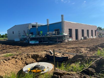 Building exterior under construction with red brick veneer, blue sheathing, and scaffolding along the wall, set on a graded dirt site with a manhole structure visible in the foreground.