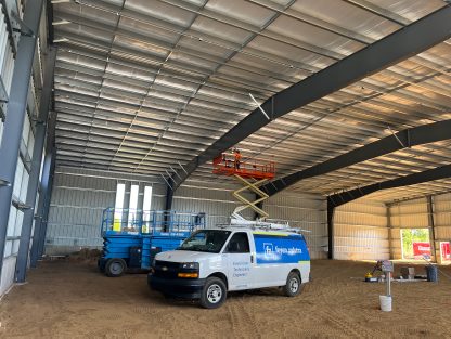 Interior of a large steel-framed building with corrugated metal roof panels, showing two scissor lifts raised near the ceiling, a work van parked inside, and a dirt floor with construction equipment and materials.