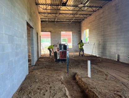 Interior of a concrete block room with a dirt floor, showing a narrow trench cut through the center, vertical utility pipes, and an open doorway leading to another unfinished area.
